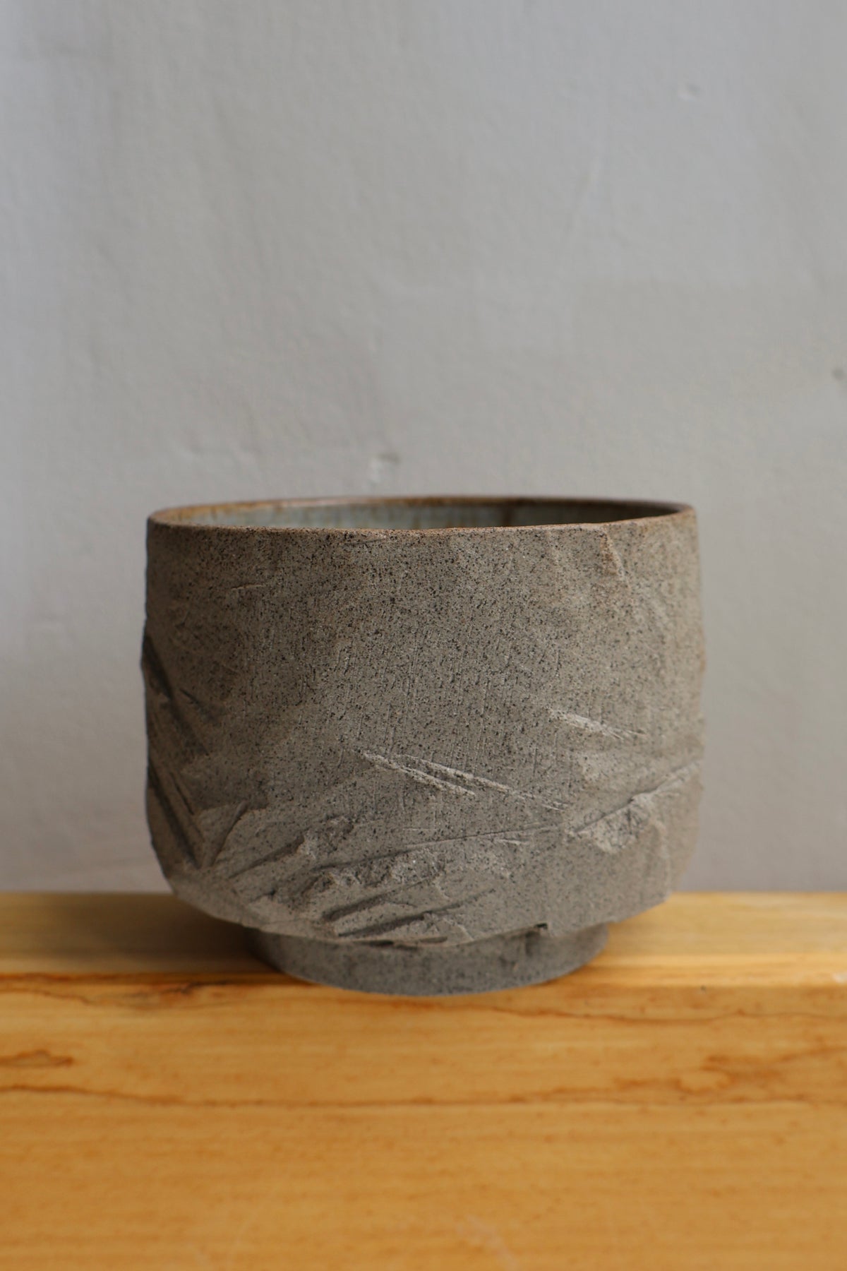 A textured gray stone or concrete planter pot with a rough, organic surface and a small base, sitting on a light wood surface against a neutral gray wall.