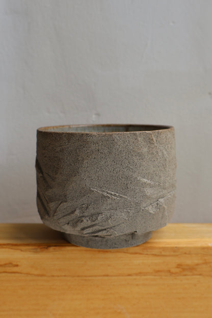 A textured gray stone or concrete planter pot with a rough, organic surface and a small base, sitting on a light wood surface against a neutral gray wall.