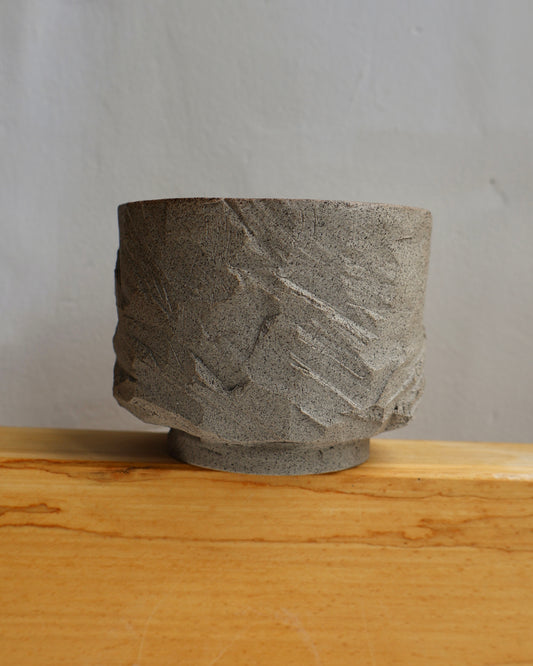 A textured gray stone bowl with a rough, hand-carved surface, resting on a light wood surface against a neutral gray background.