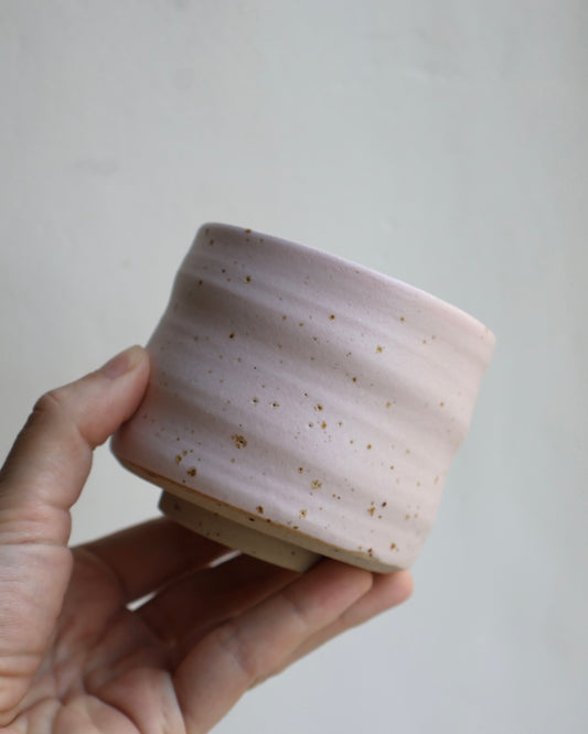 A hand holding a small handmade ceramic cup with a pale pink glaze and scattered brown speckles against a plain light gray background.