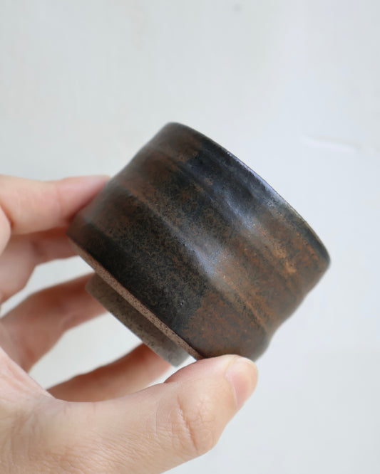 A hand holding a small handmade ceramic cup with a dark brown and black glaze, set against a plain light background.